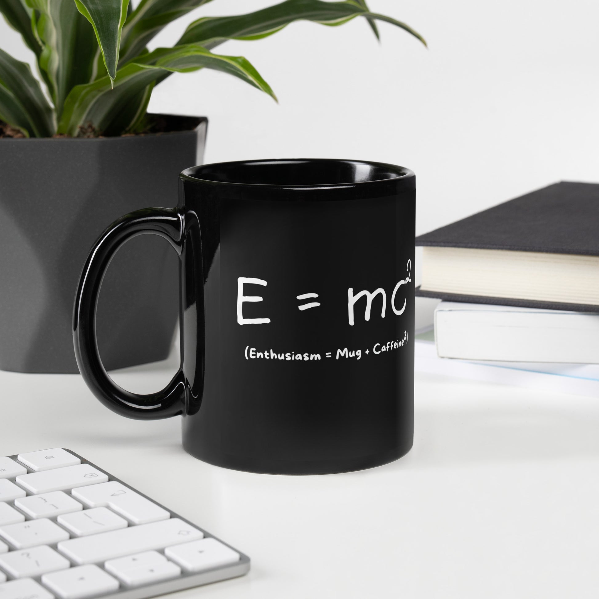 Black 11oz tech humor coffee mug featuring an ‘E=mc2’ design with handle on the left sitting on desk.