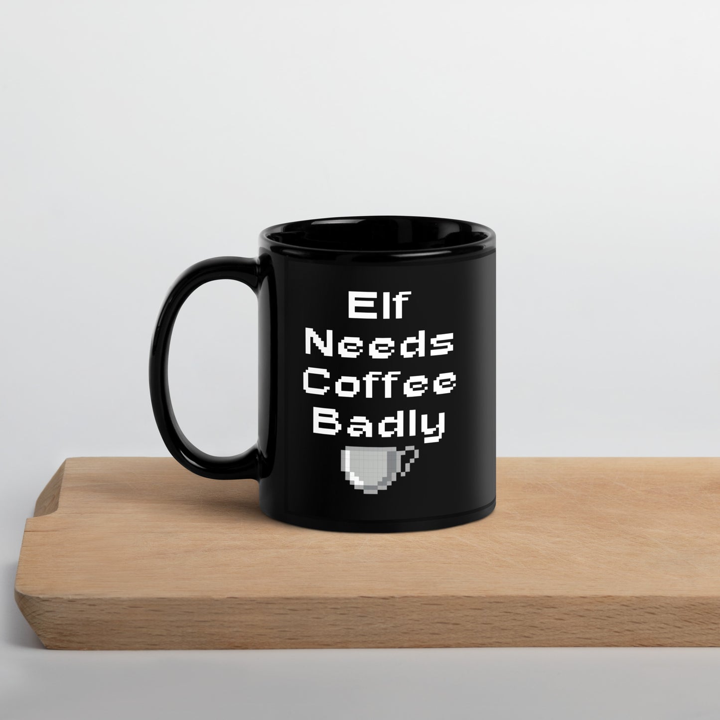 Black 11oz retro coffee mug featuring an ‘Elf Needs Coffee Badly’ video game design with handle on the left on cutting board.