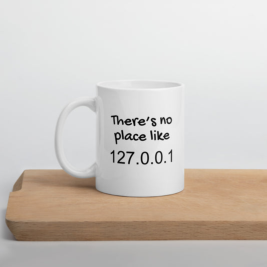 White 11oz tech humor coffee mug with the slogan ‘There's no place like 127.0.0.1’ with handle on left on cutting board.