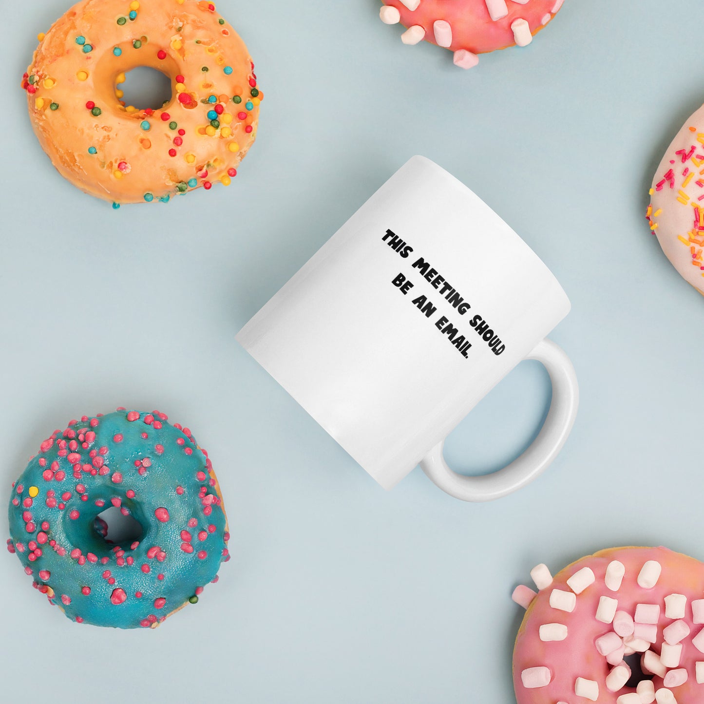 White 11oz tech humor coffee mug with the slogan ‘This Meeting Should Be An Email’ with handle on right amongst donuts.