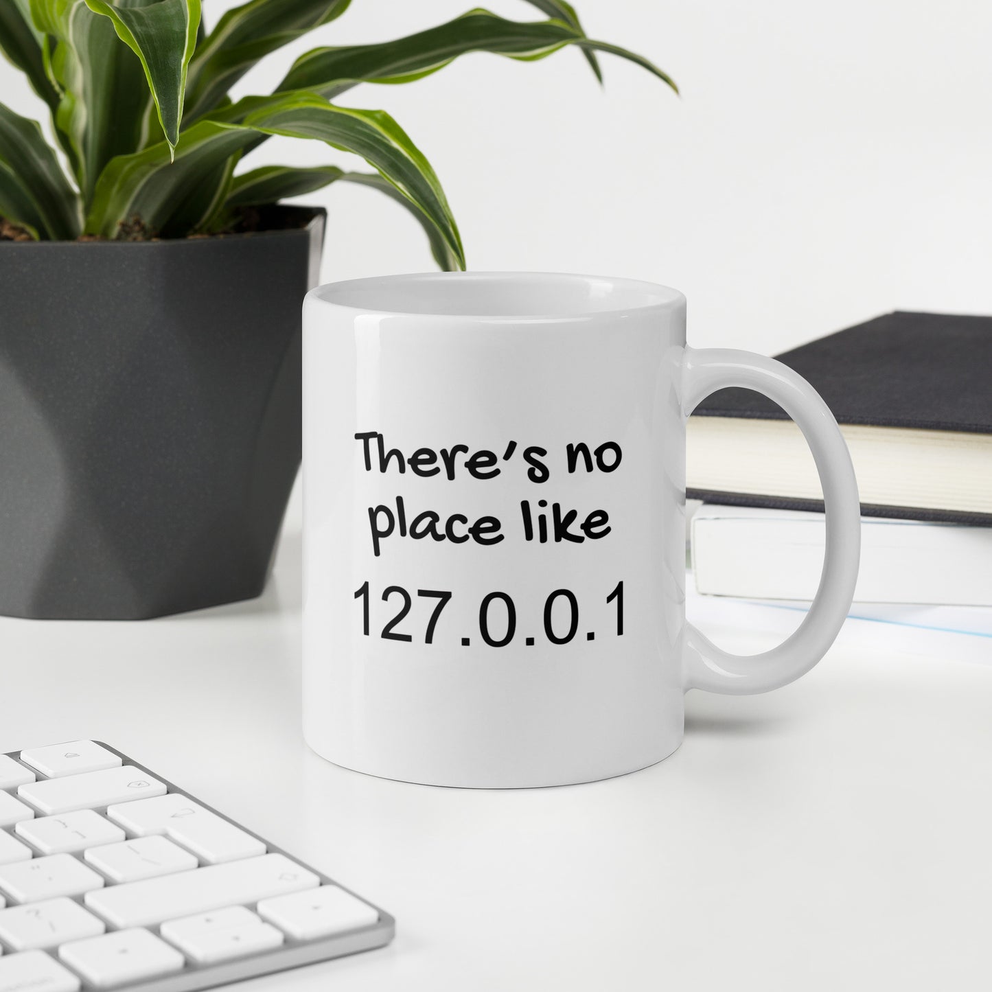 White 11oz tech humor coffee mug with the slogan ‘There's no place like 127.0.0.1’ with handle on right on office desk.