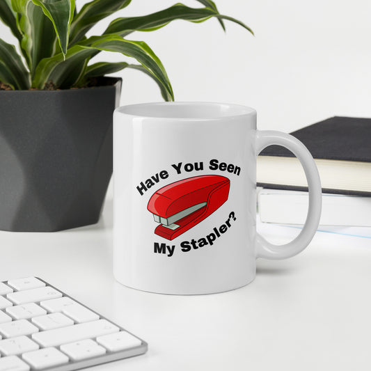 White 11oz retro tech humor coffee mug with a stapler image and slogan ‘Have you seen my stapler?’ with handle on right on desk.