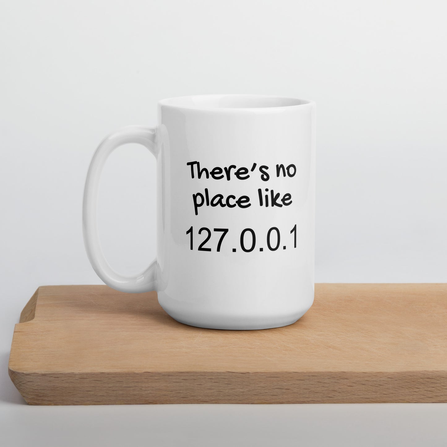 White 15oz tech humor coffee mug with the slogan ‘There's no place like 127.0.0.1’ with handle on left on cutting board.